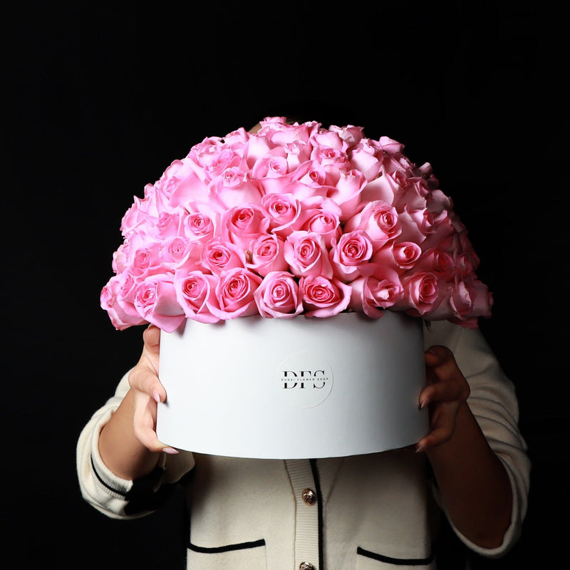 Person holding a large white box filled with pink roses against a black background